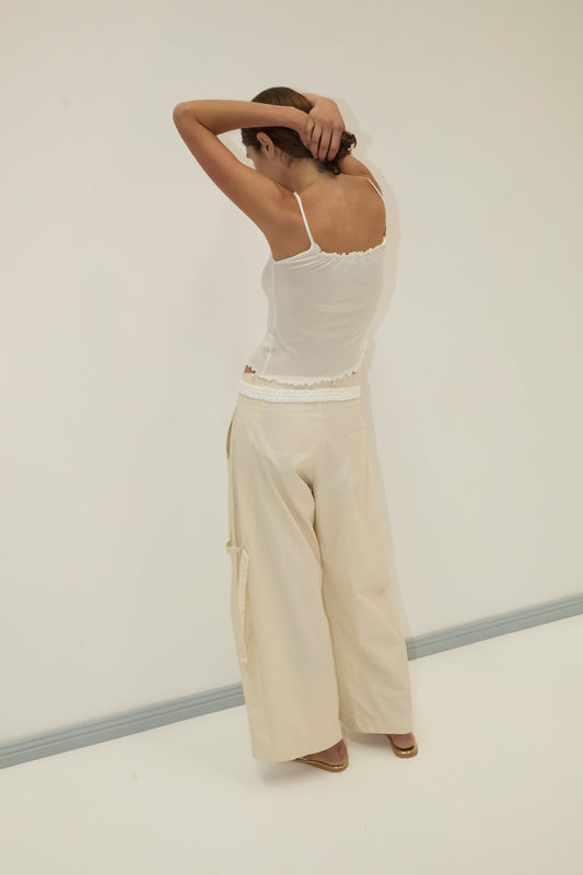 A person stands with their back to the camera, arms raised and hands behind their head. They wear a white tank top and the Tab Pant - Off White by Deiji Studios in a minimal, light-colored room.