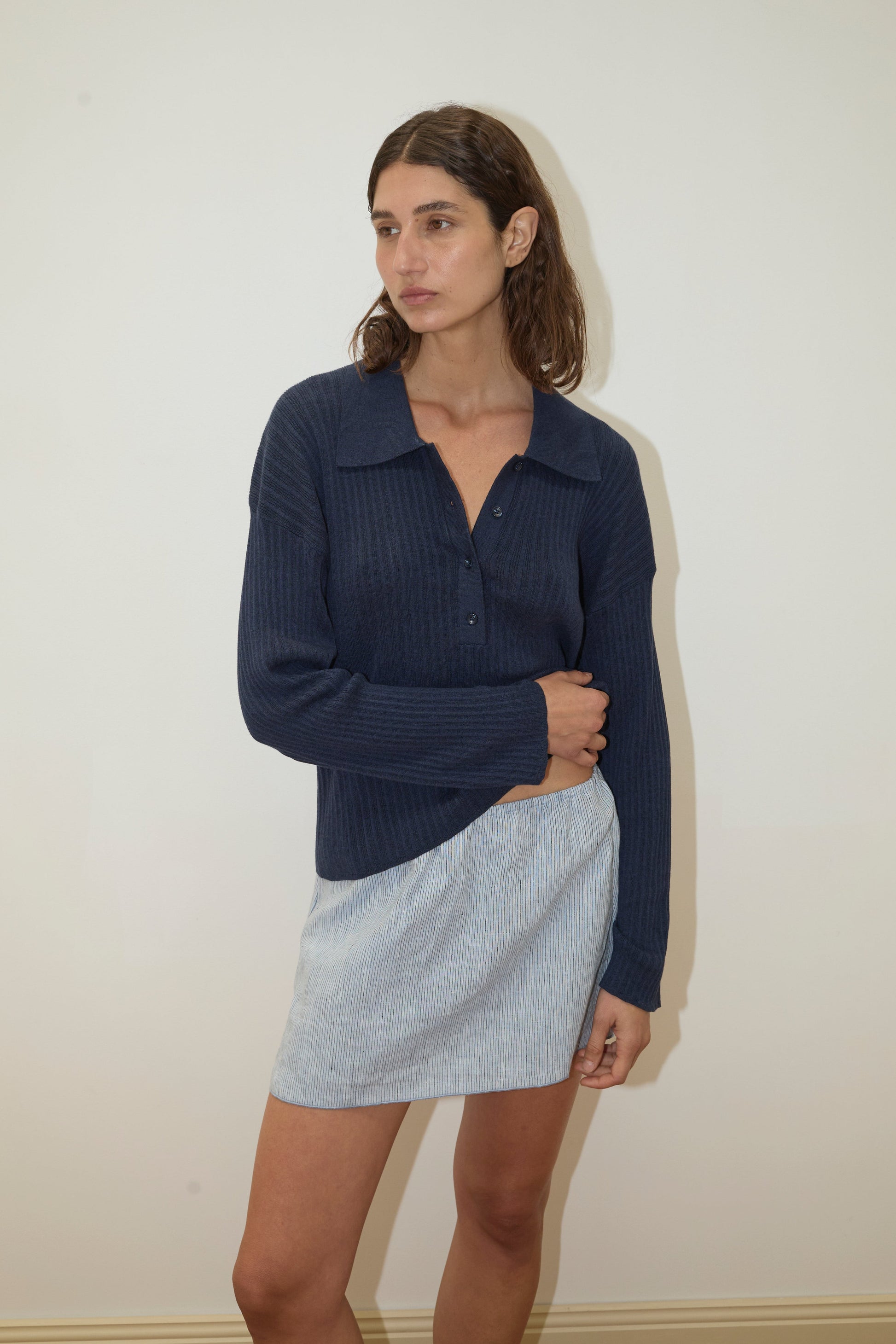 A woman with shoulder-length brown hair and a neutral expression stands against a plain light wall, wearing the Polo Knit - Dark Blue by Deiji Studios and a short, light blue skirt, with one hand holding her skirt.