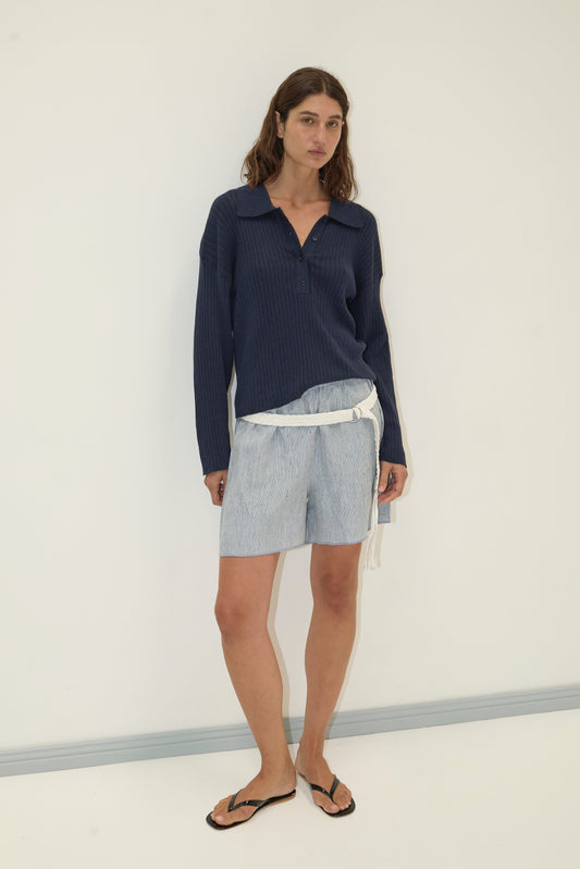 A woman with shoulder-length brown hair and a neutral expression stands against a white wall, wearing the Polo Knit - Dark Blue sweater, light blue patterned shorts with a white belt, and black sandals.