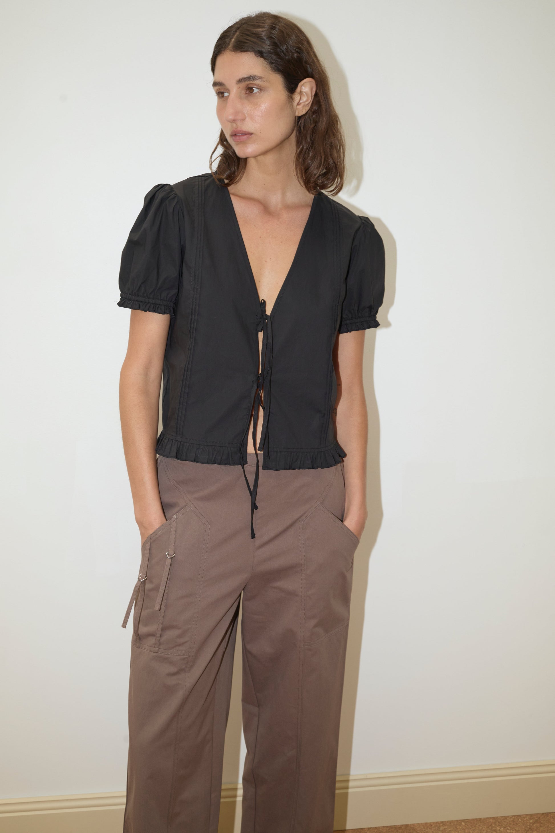A person with long brown hair stands against a white wall, wearing a Deiji Studios black short-sleeve v-neck tie-front top and loose-fitting Adjust Pocket Pant in Sawdust, with their hands in their pockets.