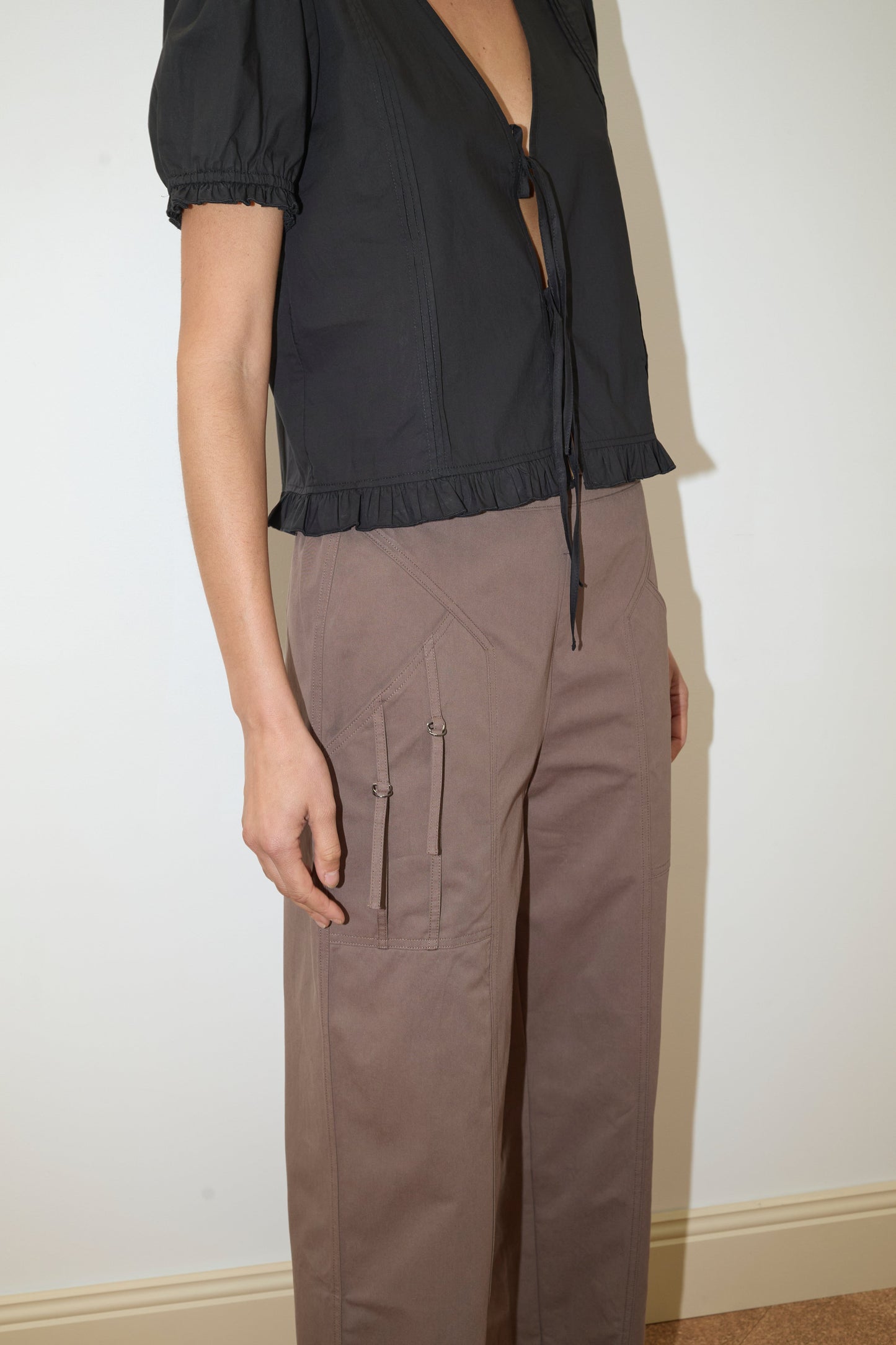 A person stands against a plain white wall wearing a Deiji Studios short-sleeved black blouse with a deep V-neck and Adjust Pocket Pant – Sawdust. Their face is not visible.