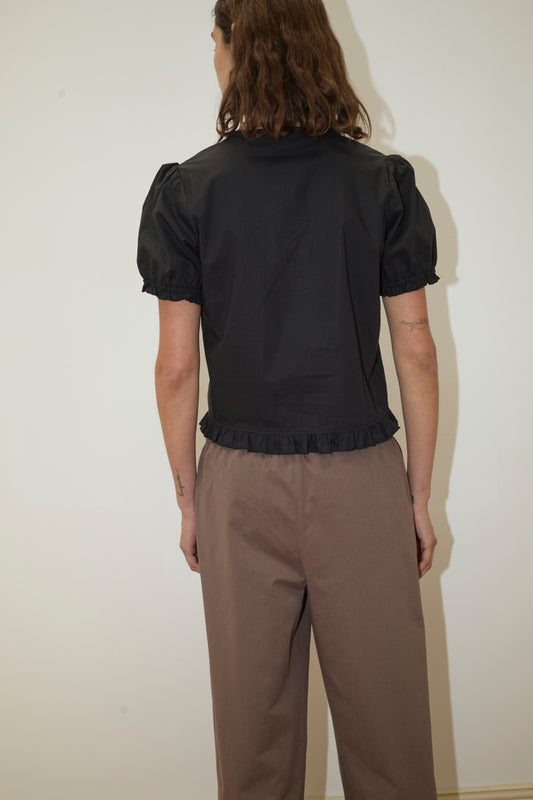 A person with wavy brown hair, facing away, wears a short-sleeve black ruffle blouse and high-waisted Adjust Pocket Pant - Sawdust by Deiji Studios in organic cotton, standing against a plain light-colored wall.