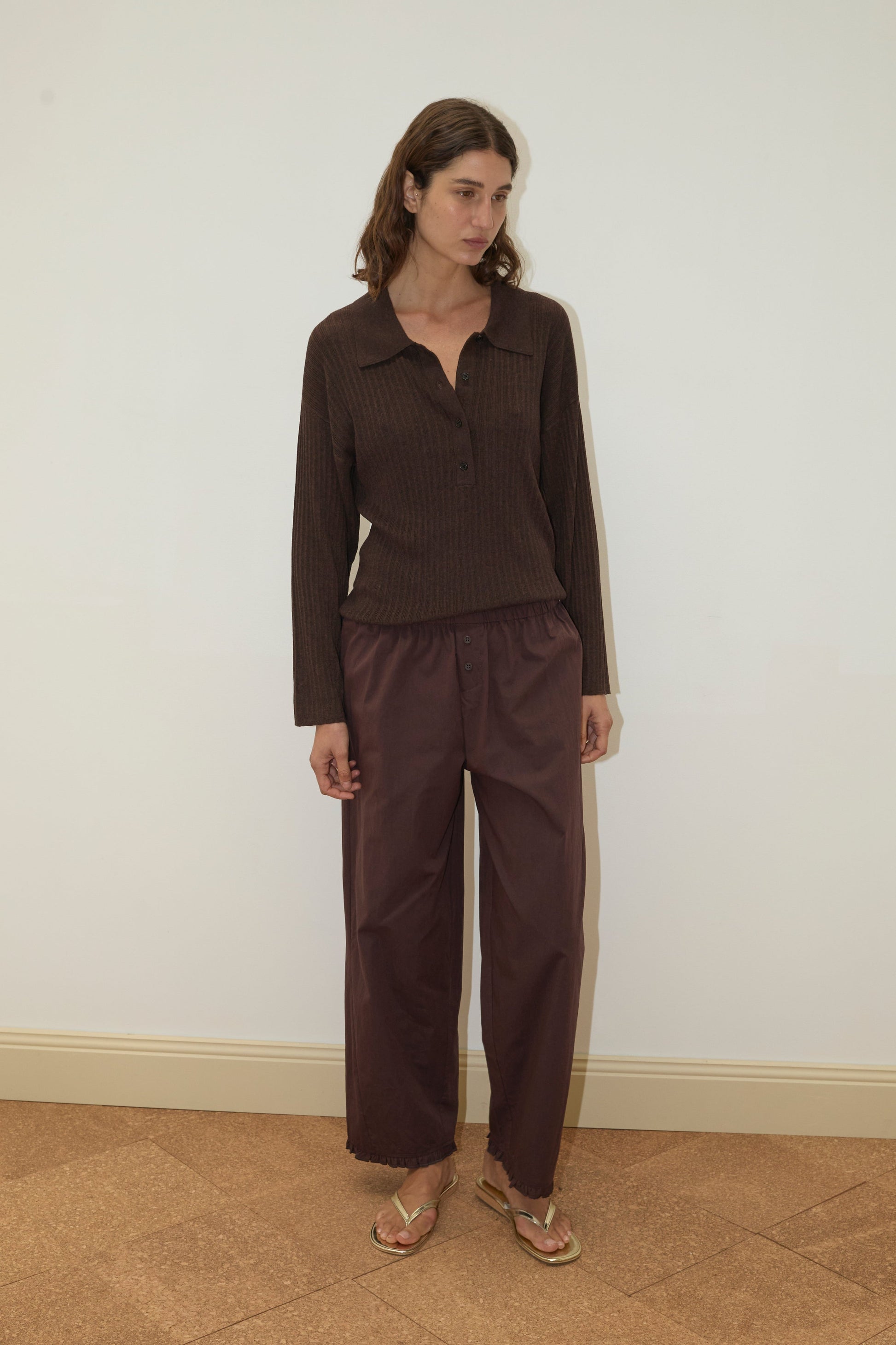 A woman with wavy brown hair stands against a plain light wall, wearing the Polo Knit - Dark Almond long-sleeve collared shirt, loose maroon pants, and sandals, looking slightly to the side.