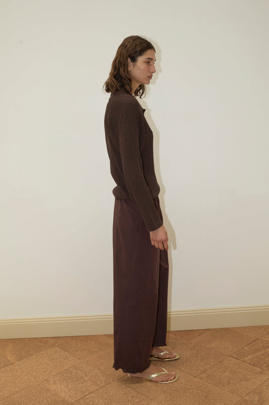 A person with shoulder-length hair stands sideways against a plain white wall, wearing a Deiji Studios Polo Knit in Dark Almond, loose brown organic cotton pants, and sandals on a tan tiled floor.