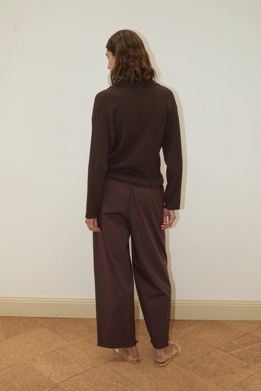 A person with wavy brown hair stands facing a white wall, wearing the Polo Knit - Dark Almond sweater from Deiji Studios, paired with loose maroon pants and beige sandals on a tan tiled floor.
