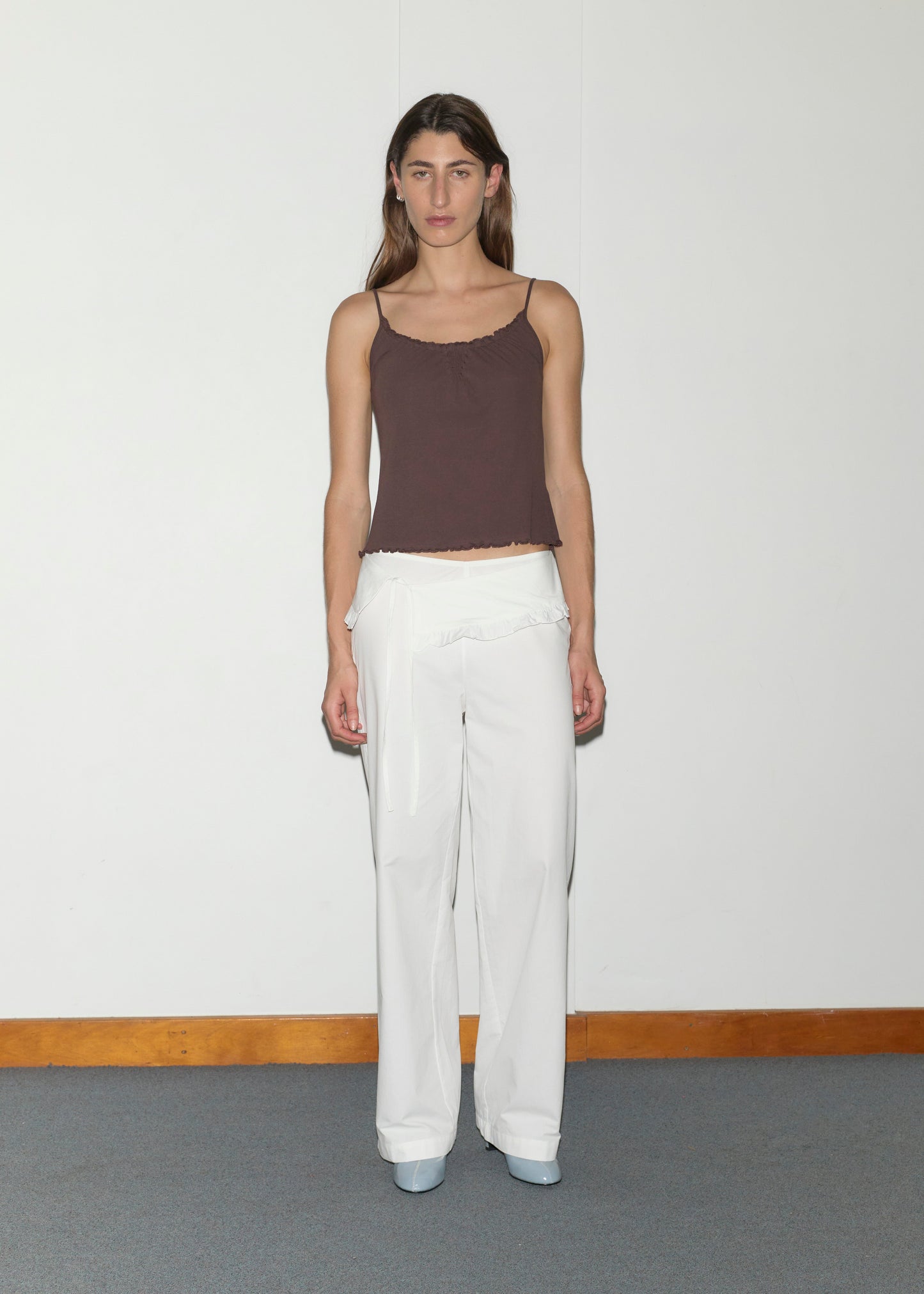 A woman with long brown hair stands against a plain white wall, wearing a brown spaghetti strap top, pale blue shoes, and the Flounce Belt Pant in white with ruffle detail. She faces the camera with a neutral expression.