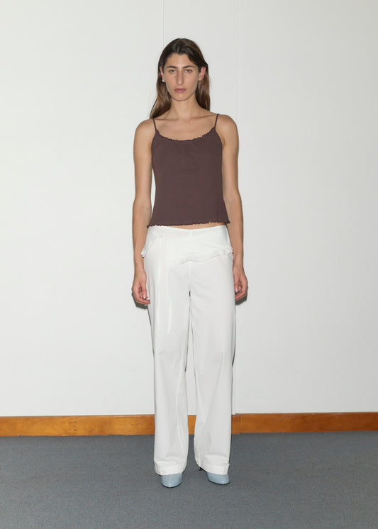 A woman with long brown hair stands against a plain white wall, wearing a brown spaghetti strap top, pale blue shoes, and the Flounce Belt Pant in white with ruffle detail. She faces the camera with a neutral expression.