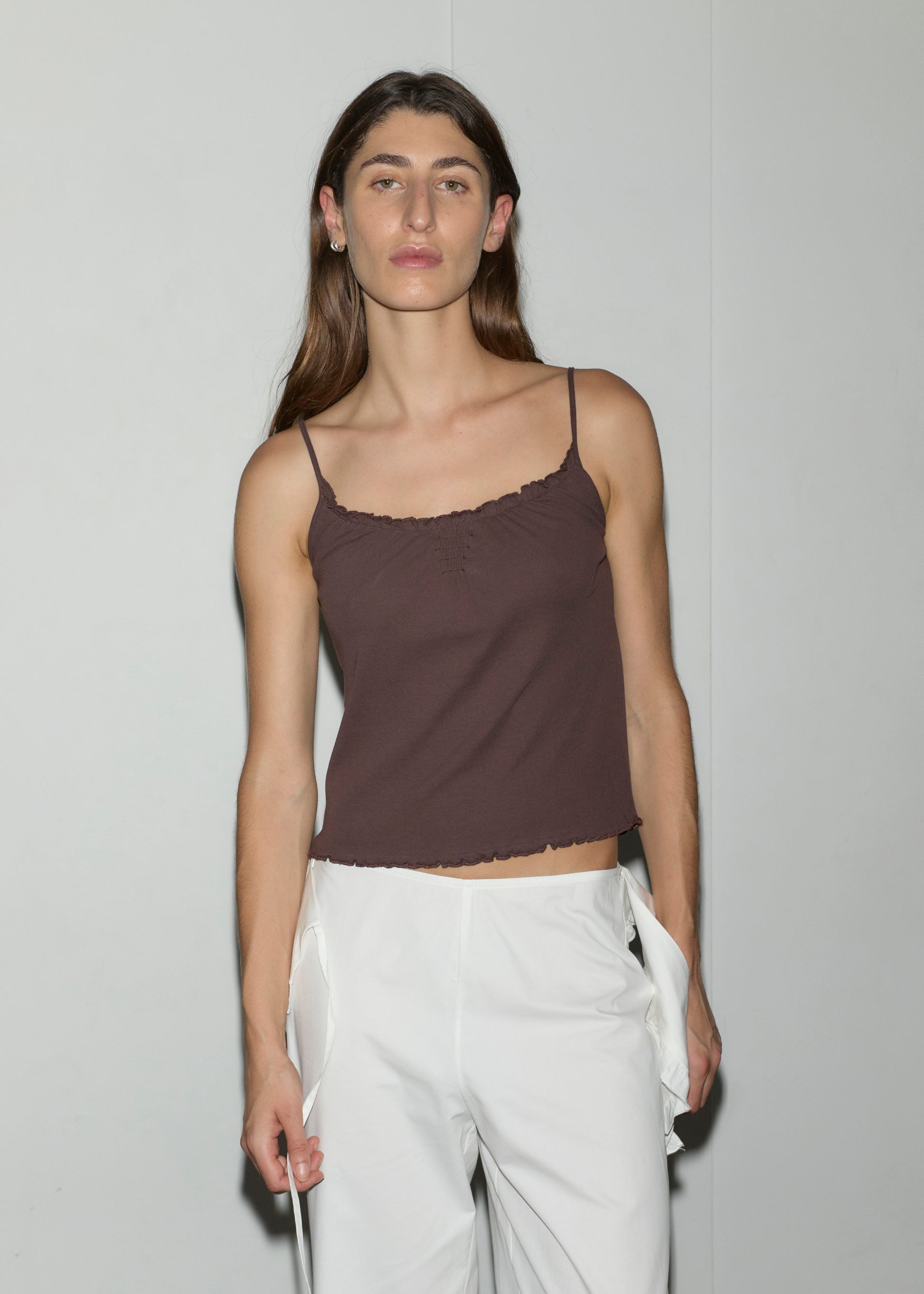 A person with long brown hair stands against a plain light wall, wearing the Cotton Flounce Tank in brown and white pants, looking directly at the camera with a neutral expression.