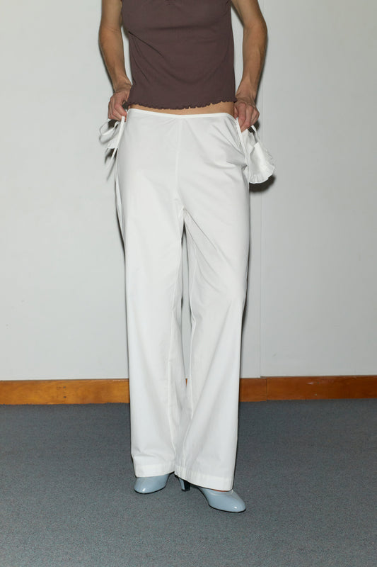 A person stands indoors, head out of frame, holding out the waistband of loose white Flounce Belt Pant by Deiji Studios to show the fit. Styled with a sleeveless brown top and light blue heeled shoes.