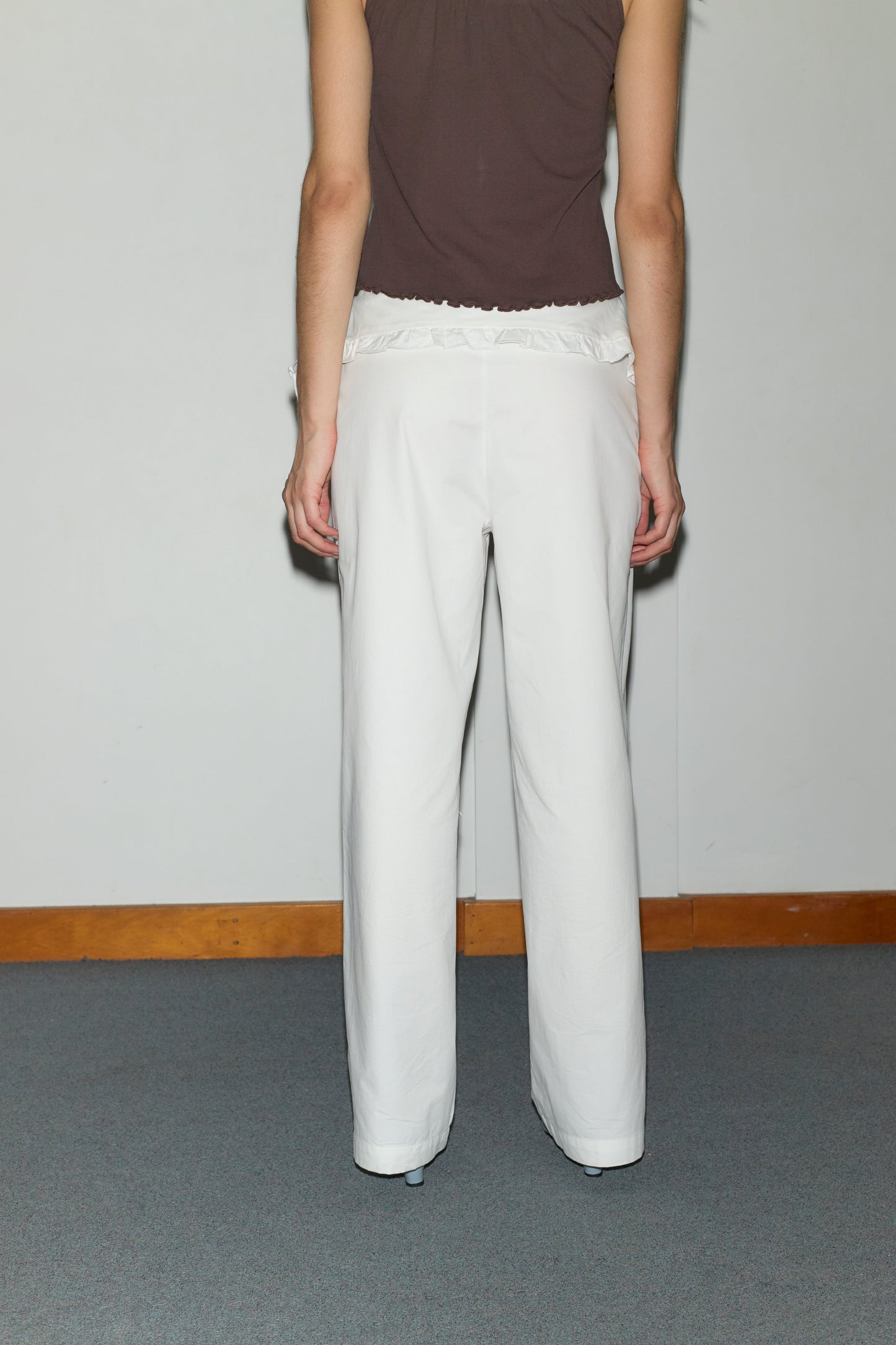 A person stands facing away from the camera, wearing a sleeveless brown top and the Flounce Belt Pant - White against a plain wall with wooden trim and a gray carpeted floor.