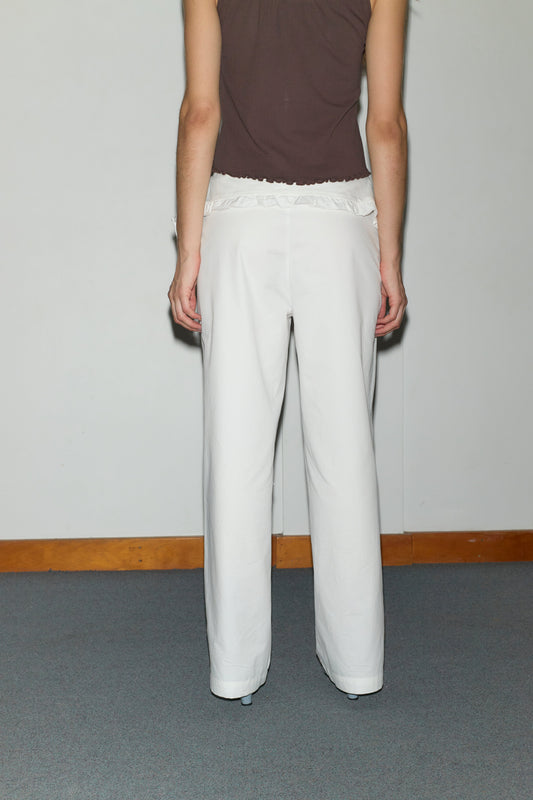 A person stands facing a wall, shown from the back, wearing a sleeveless dark brown top and Deiji Studios’ Flounce Belt Pant in white. The indoor setting features a gray carpet and wooden baseboard along the wall.
