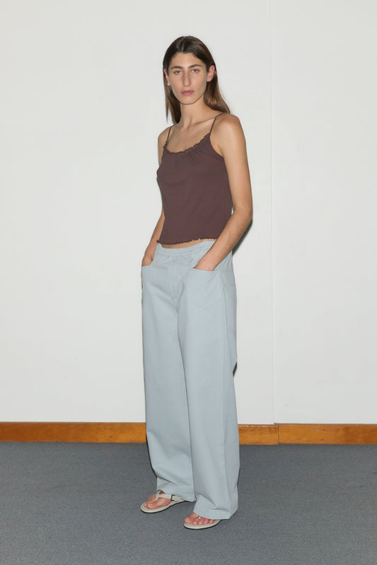 A woman with long brown hair stands against a white wall, wearing a brown spaghetti-strap top, the Square Pocket Pant - Blue by Deiji Studios with square pockets, and sandals. Her hands are in her pockets.