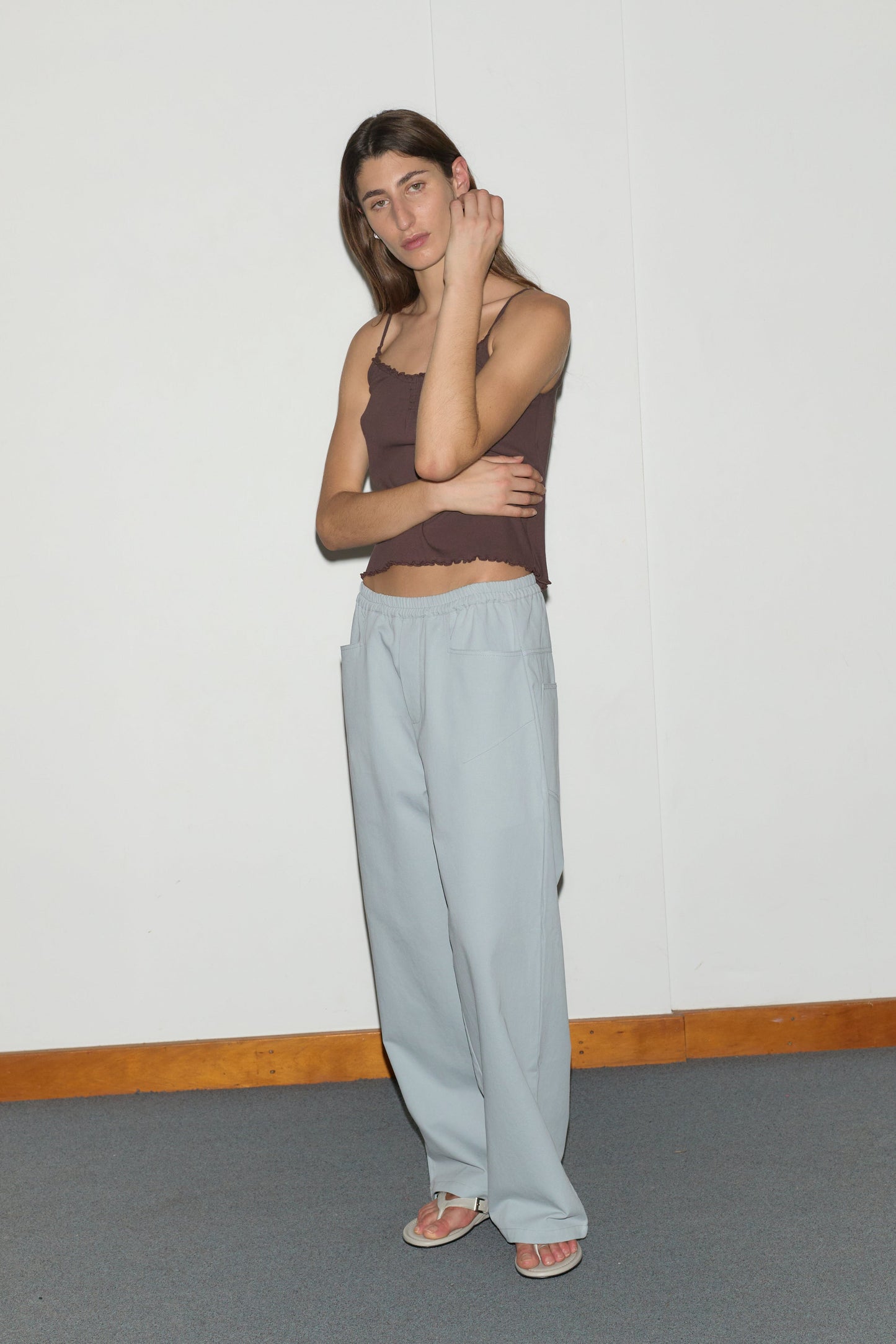 A person stands against a plain white wall, wearing a brown sleeveless top and Square Pocket Pant in blue with sandals. They have long hair and rest one arm across their body while touching their neck.