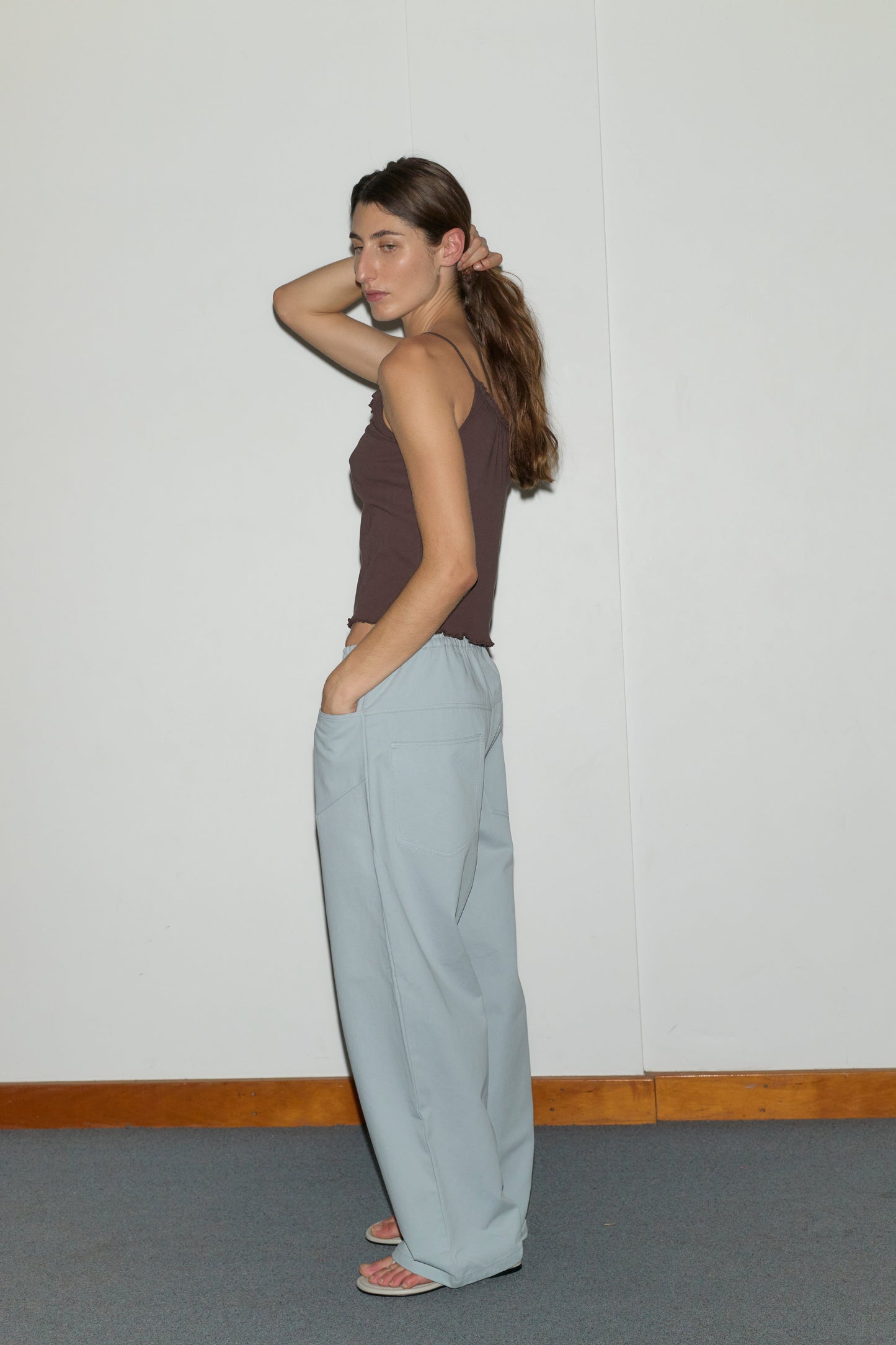 A woman stands sideways against a white wall, wearing the Square Pocket Pant - Blue with wide, straight legs, a brown sleeveless top, and sandals. She touches her hair with one hand and looks relaxed.