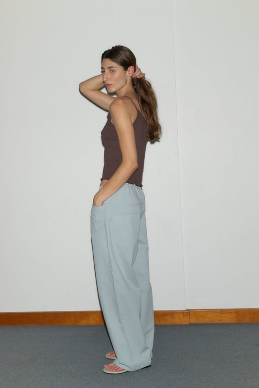 A woman stands sideways against a plain white wall, wearing a brown sleeveless top and Deiji Studios' Square Pocket Pant - Blue. She appears relaxed in sandals with one hand raised to her hair.