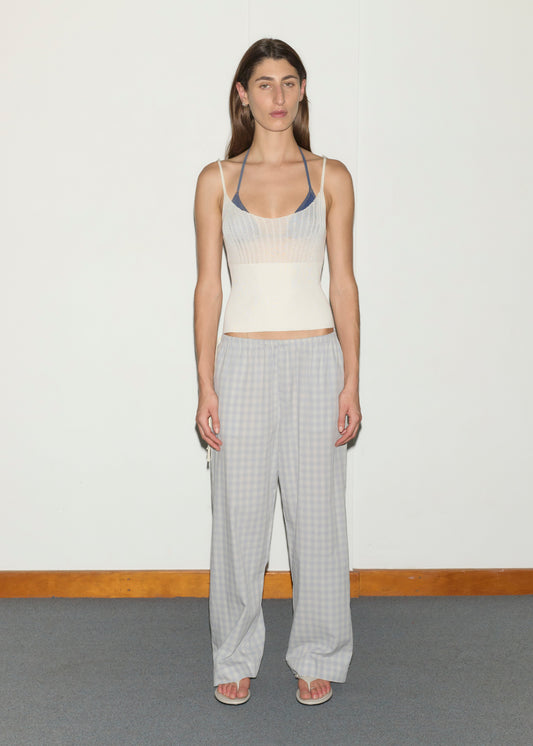 A person stands against a white wall wearing a white knit tank top over a blue bra, Side Tie Pant - Blue Check by Deiji Studios, and white flip-flops. The floor is carpeted with a wooden baseboard.
