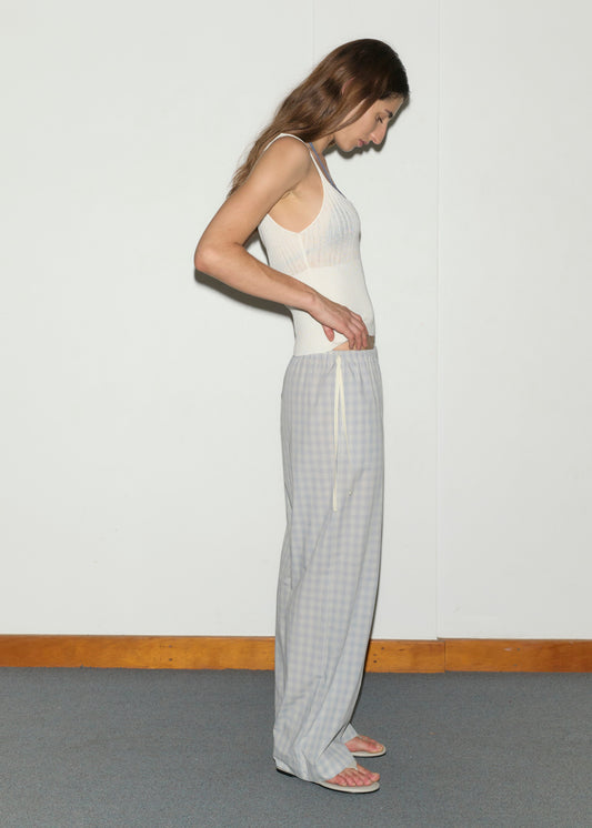 A woman in profile stands against a plain wall, wearing a white tank top, sandals, and the Side Tie Pant - Blue Check by Deiji Studios. She looks down with one hand resting on her waistband.