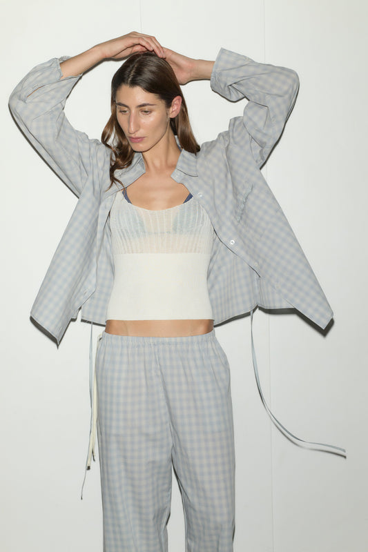 A woman stands against a white wall wearing the Side Tie Pant - Blue Check over a textured white camisole, arms raised with hands on her head, looking slightly downward.