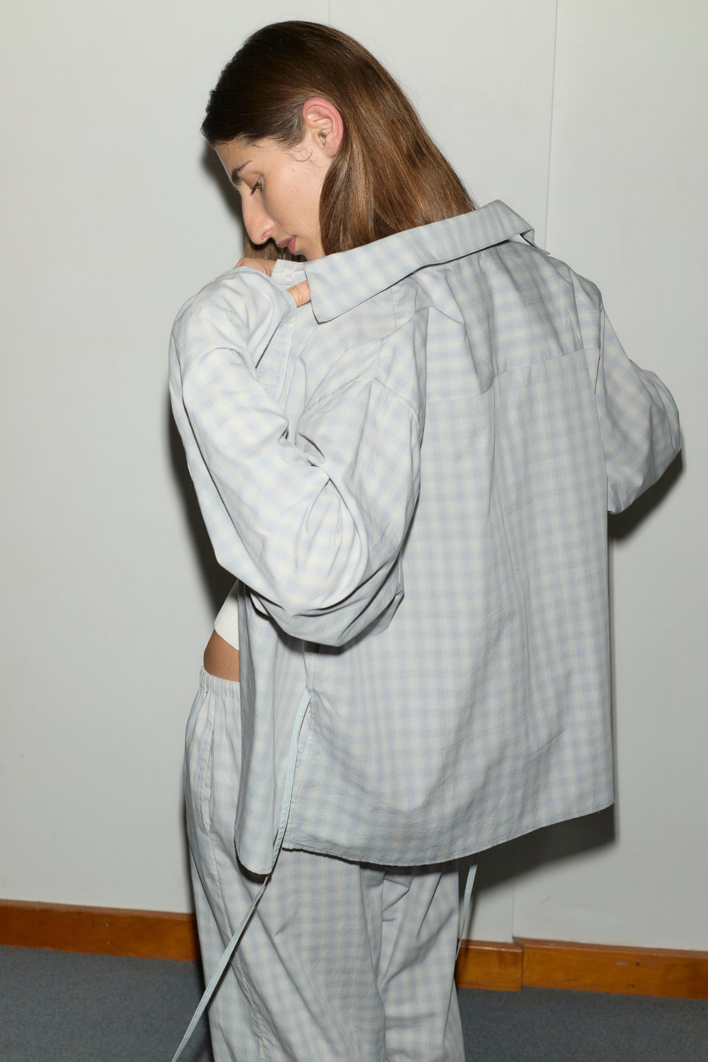 A person with long brown hair stands sideways, looking down and adjusting the collar of the Day Shirt - Blue Check. The shirt has a boxy fit and light blue check pattern. Part of their side is visible against a plain background with a wooden trim.