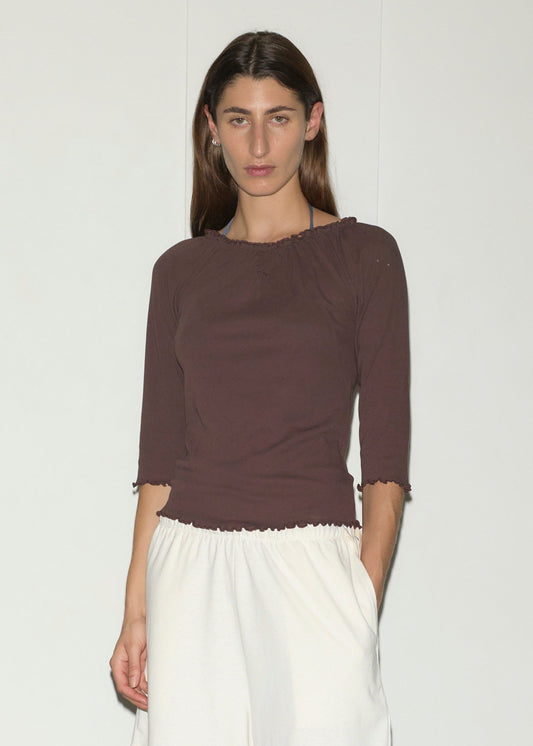 A person with long brown hair wears the Cotton Flounce 3/4 Sleeve - Brown by Deiji Studios and loose white pants, standing against a plain white background with one hand in their pocket.