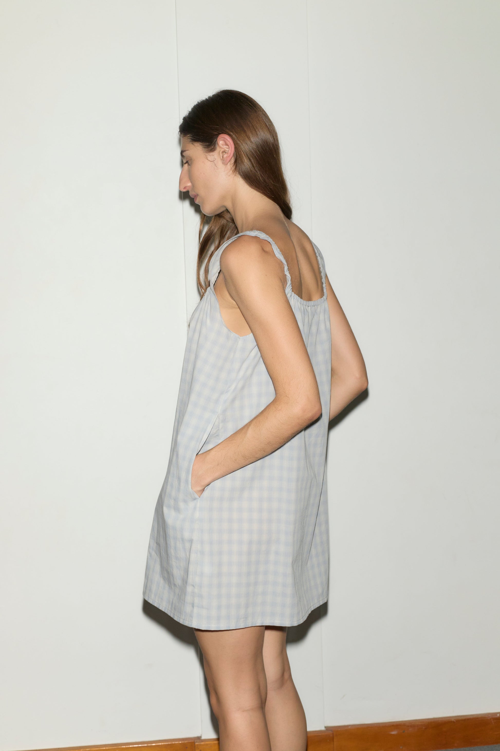 A person with long brown hair stands sideways against a plain white wall and wooden floor, wearing the bow dress - blue check, a mini-length dress featuring pockets.