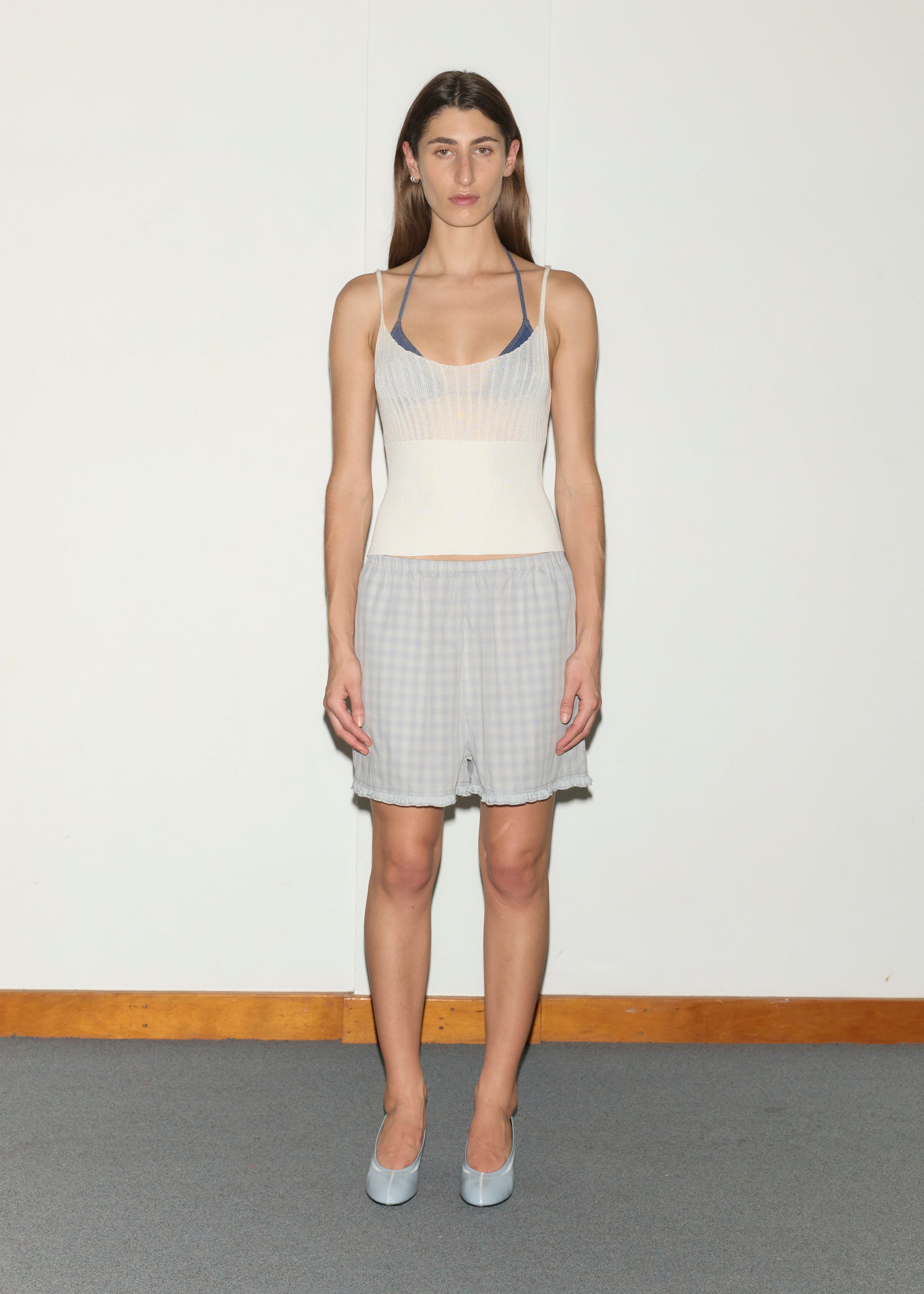 A woman stands indoors on gray carpet against a white wall, wearing the Simple Short - Blue Check, a cream sleeveless top over a blue bralette, and light blue heels, looking directly at the camera.