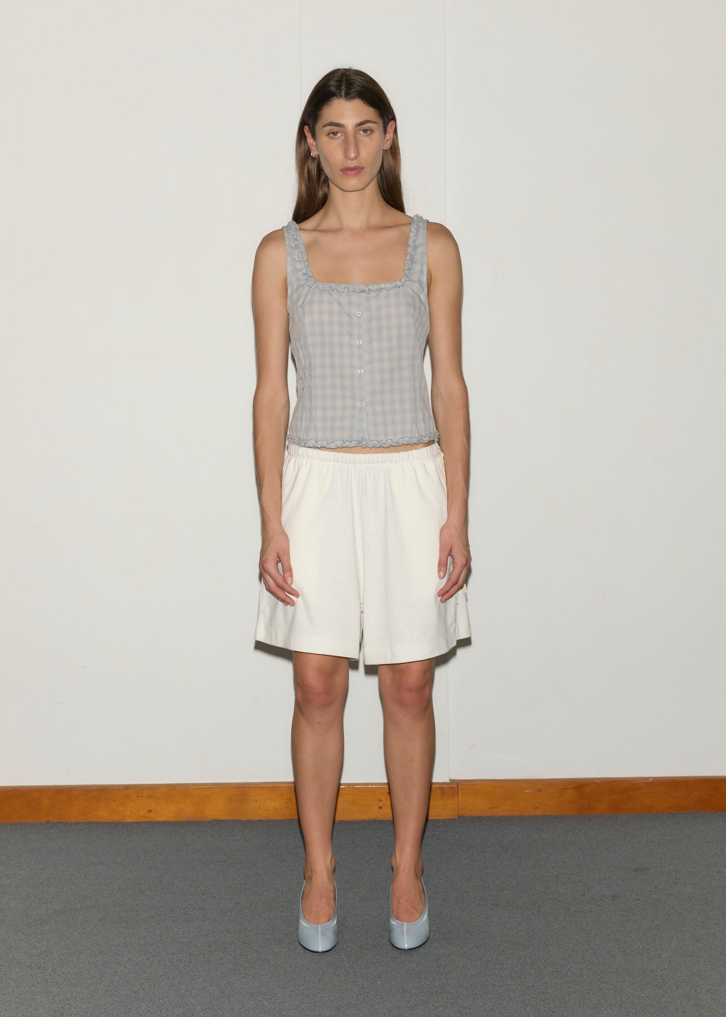 A person with long brown hair stands facing forward against a white wall, wearing a Deiji Studios light blue cotton poplin sleeveless top, Relax Short - Off White, blue shoes, with a wooden baseboard and gray floor in the background.