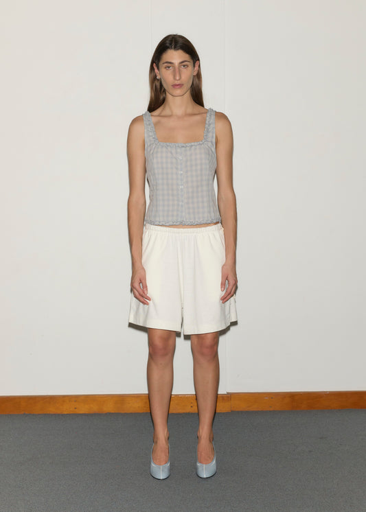 A woman with long brown hair stands against a plain white wall, wearing the Relax Short in Off White with an elastic waist and a sleeveless, light blue checkered top. She pairs the outfit with light blue shoes. The floor is carpeted; the wall has wooden trim.