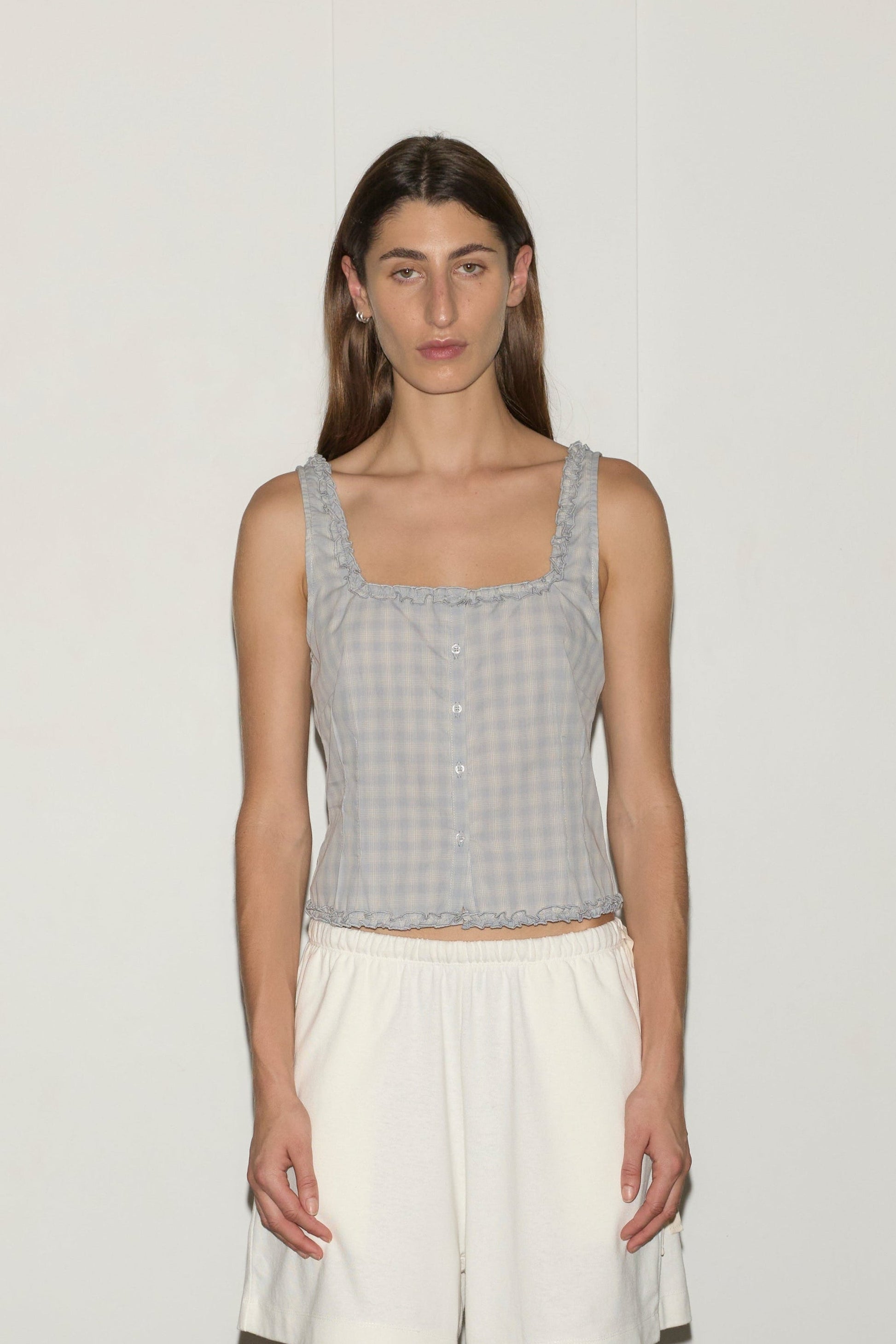 A person with long brown hair stands against a plain white background, wearing the Button Frill Top - Blue Check from Deiji Studios and white shorts. They face the camera with a neutral expression.