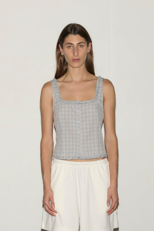 A person with long brown hair stands against a plain white background, wearing the Button Frill Top - Blue Check from Deiji Studios and white shorts. They face the camera with a neutral expression.