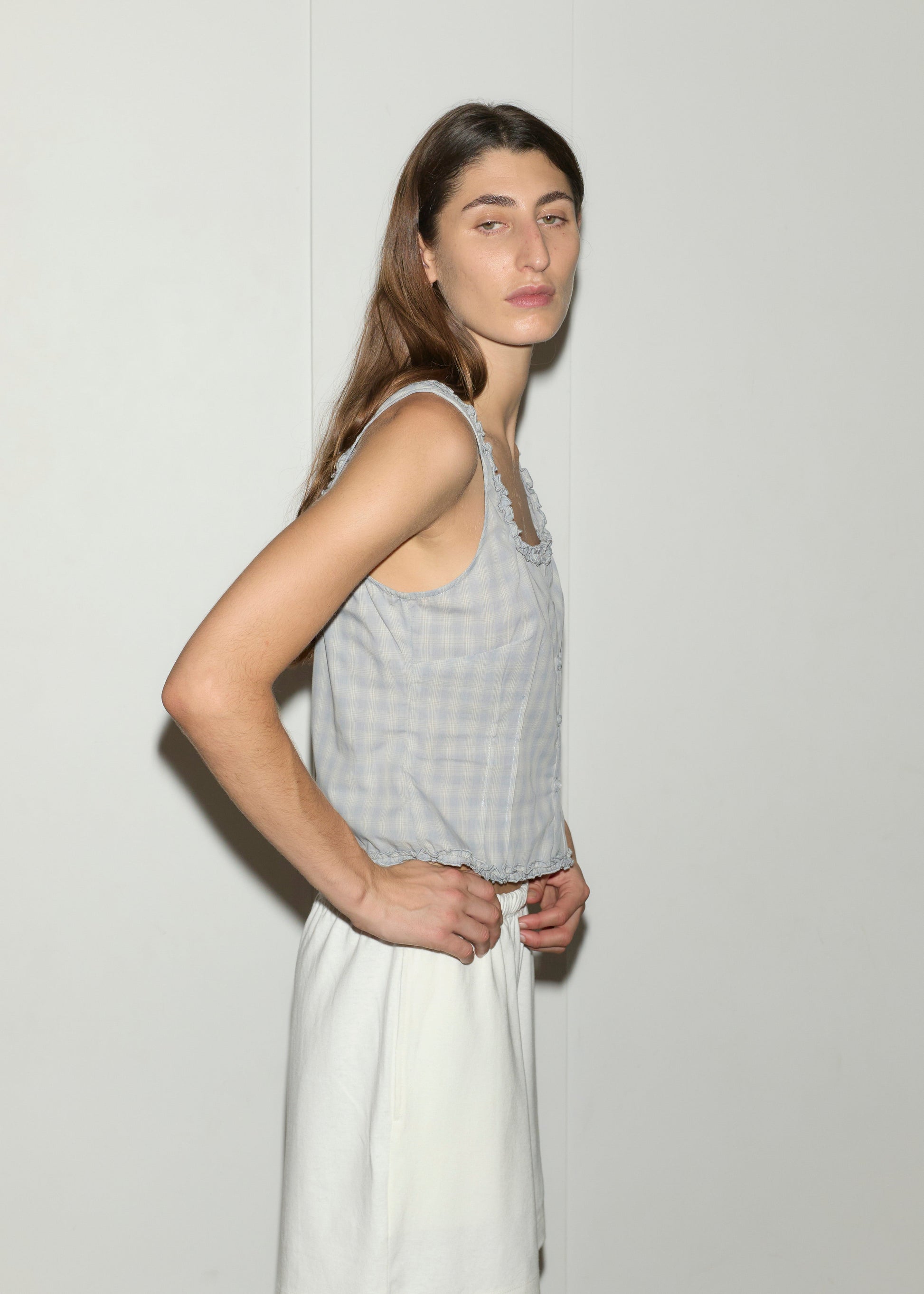 A person with long brown hair wears the Button Frill Top - Blue Check by Deiji Studios and a white skirt, standing in front of a plain white wall and looking slightly to the side with a neutral expression.
