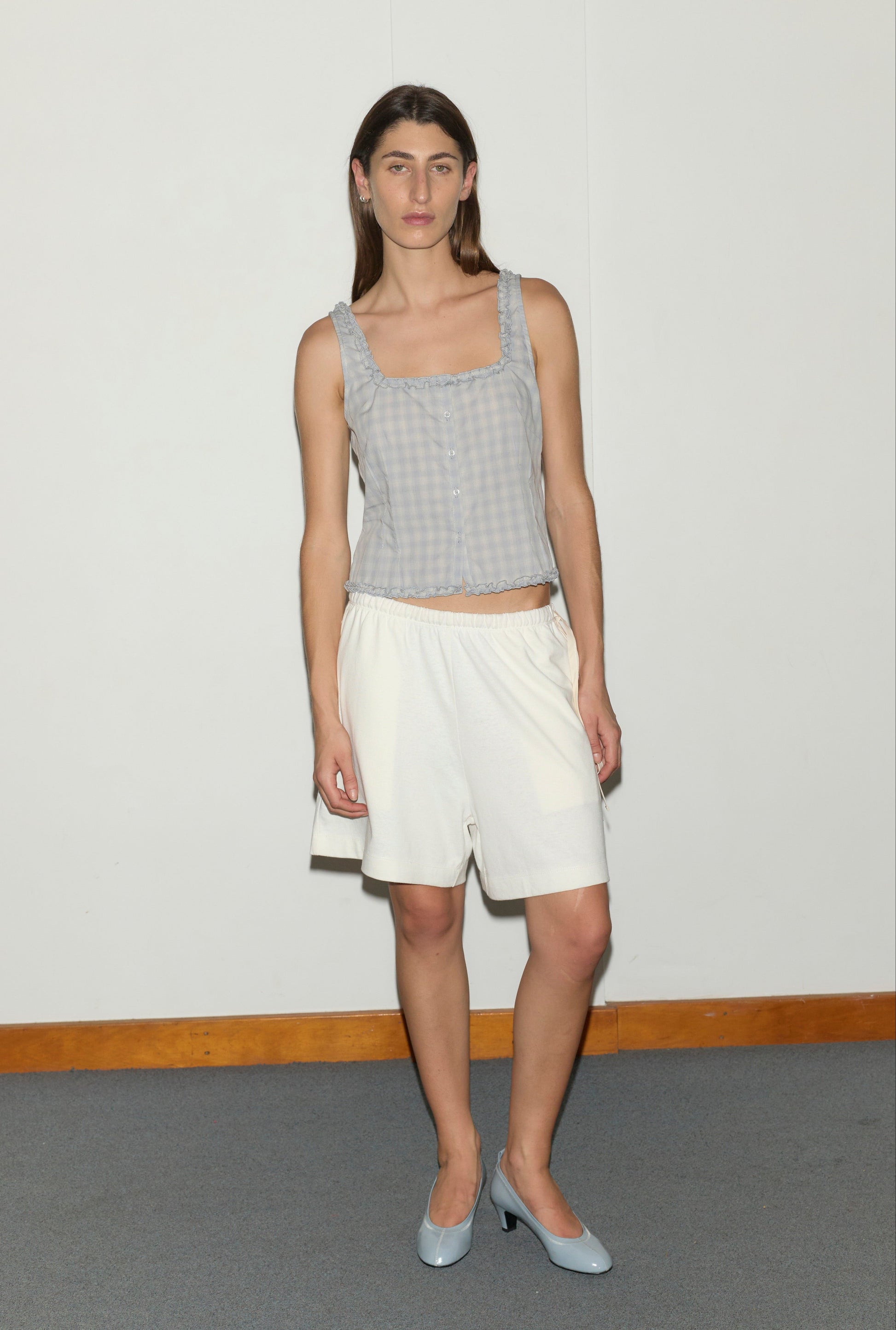 A person with long brown hair stands indoors against a white wall, wearing the Button Frill Top - Blue Check from Deiji Studios, loose white shorts, and light blue heels. They appear relaxed with a neutral expression.