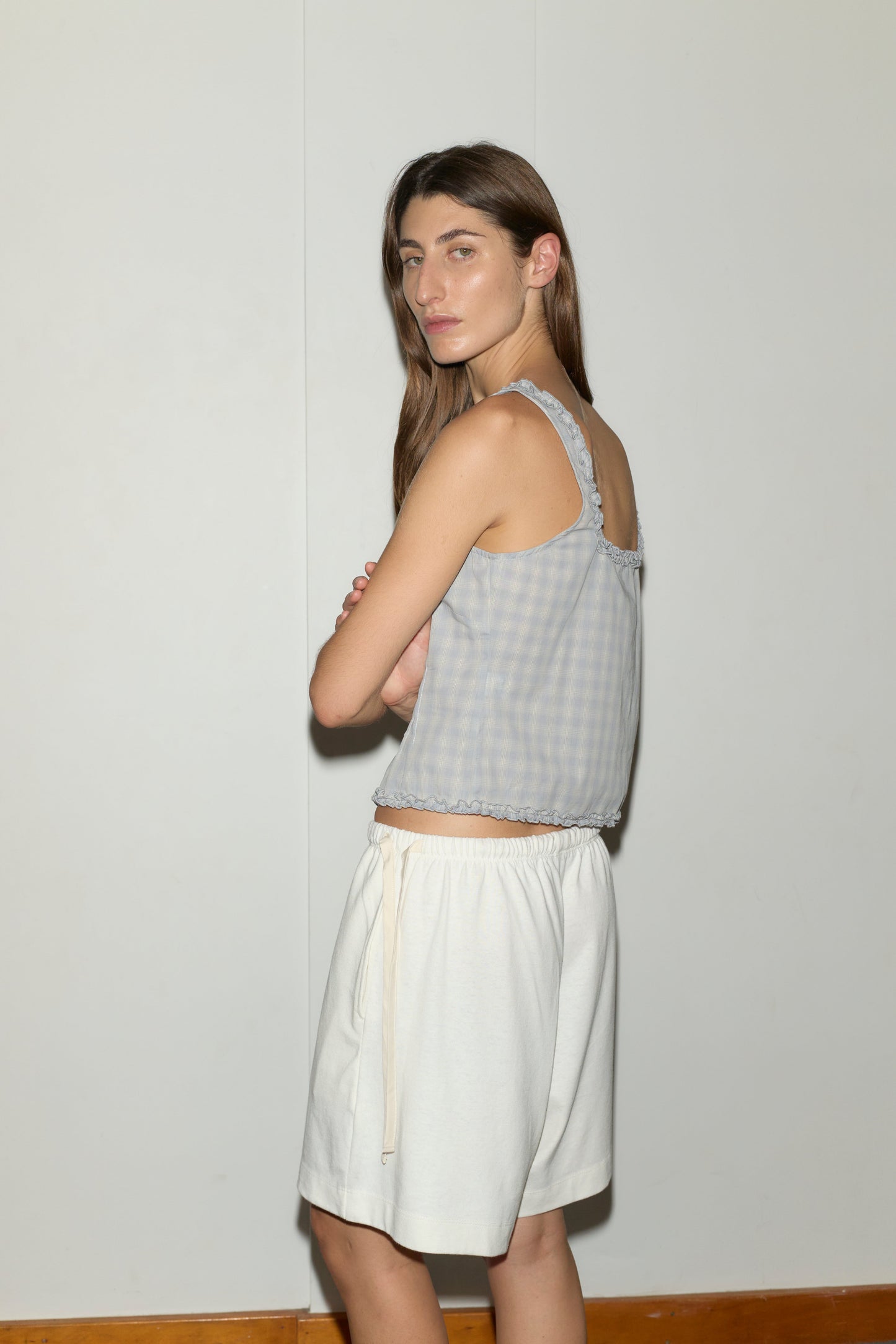 A woman with long brown hair wears the Button Frill Top - Blue Check from Deiji Studios and loose white shorts, standing with arms crossed and looking over her shoulder against a plain light wall.