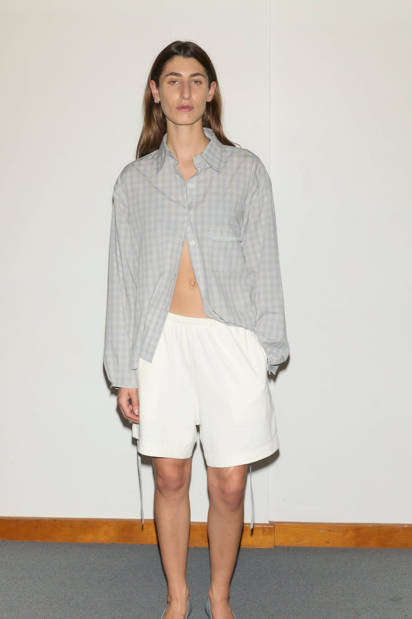 A person stands indoors against a plain white wall, wearing Deiji Studios' loose-fitting, light gray cotton poplin checkered shirt unbuttoned at the bottom and the Relax Short - Off White. They have long brown hair and a neutral expression.