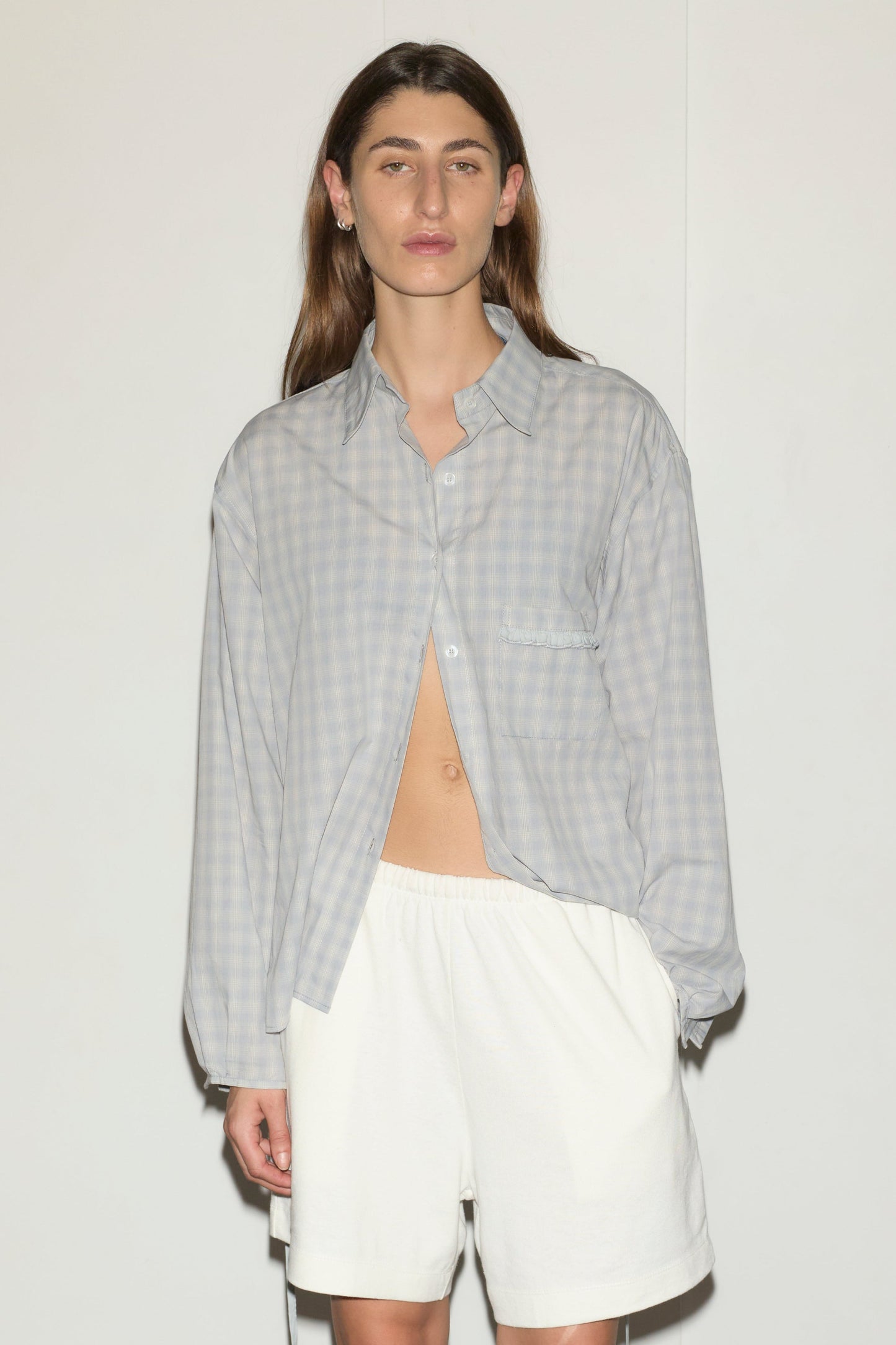 A person with long brown hair wears a light gray checked day shirt, partially unbuttoned, and loose white shorts, standing against a plain white background.