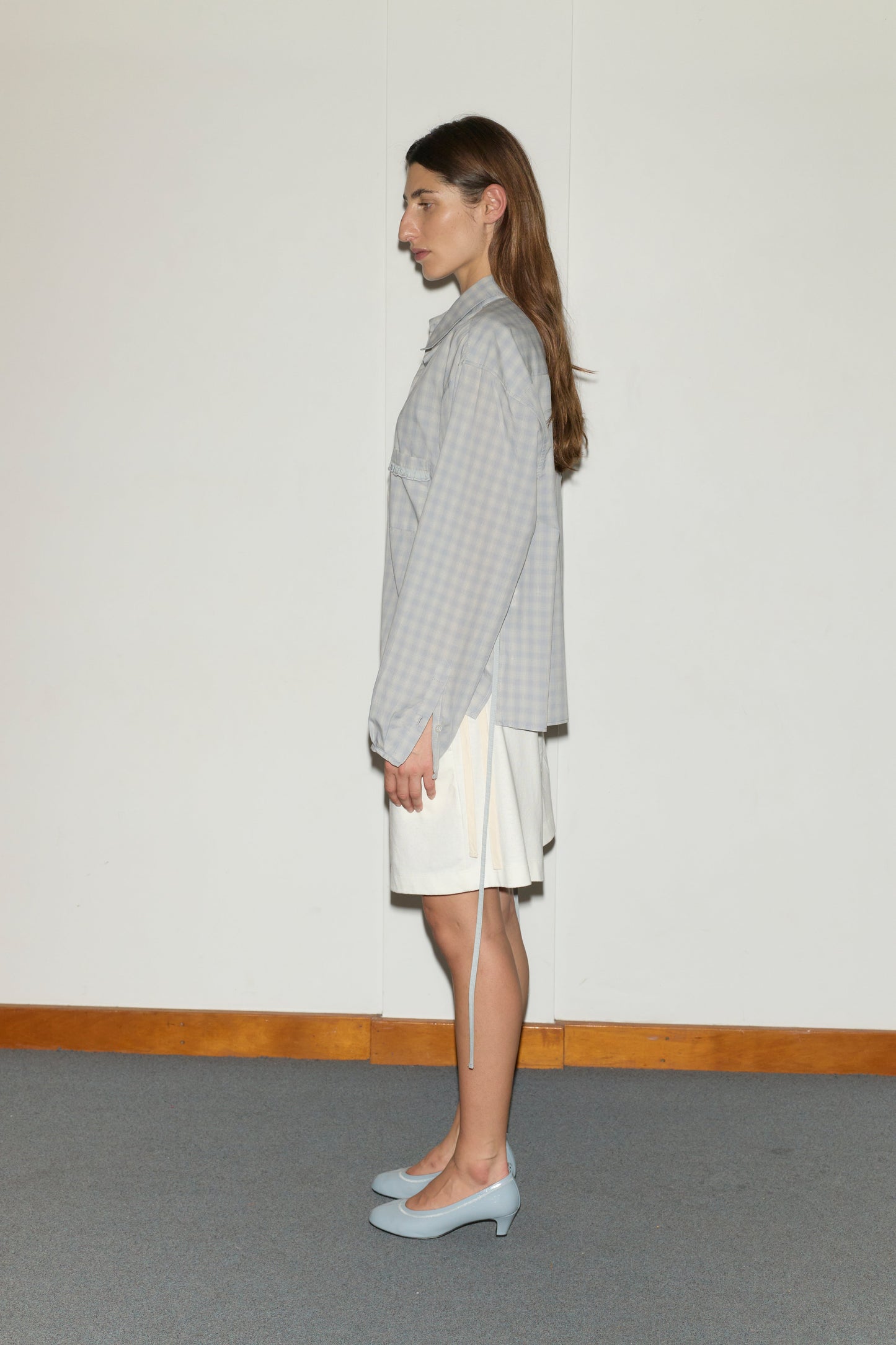 A woman with long brown hair stands in profile against a white wall, wearing the Day Shirt - Blue Check, a white skirt, and light blue heels on a carpeted floor edged with a wooden baseboard.