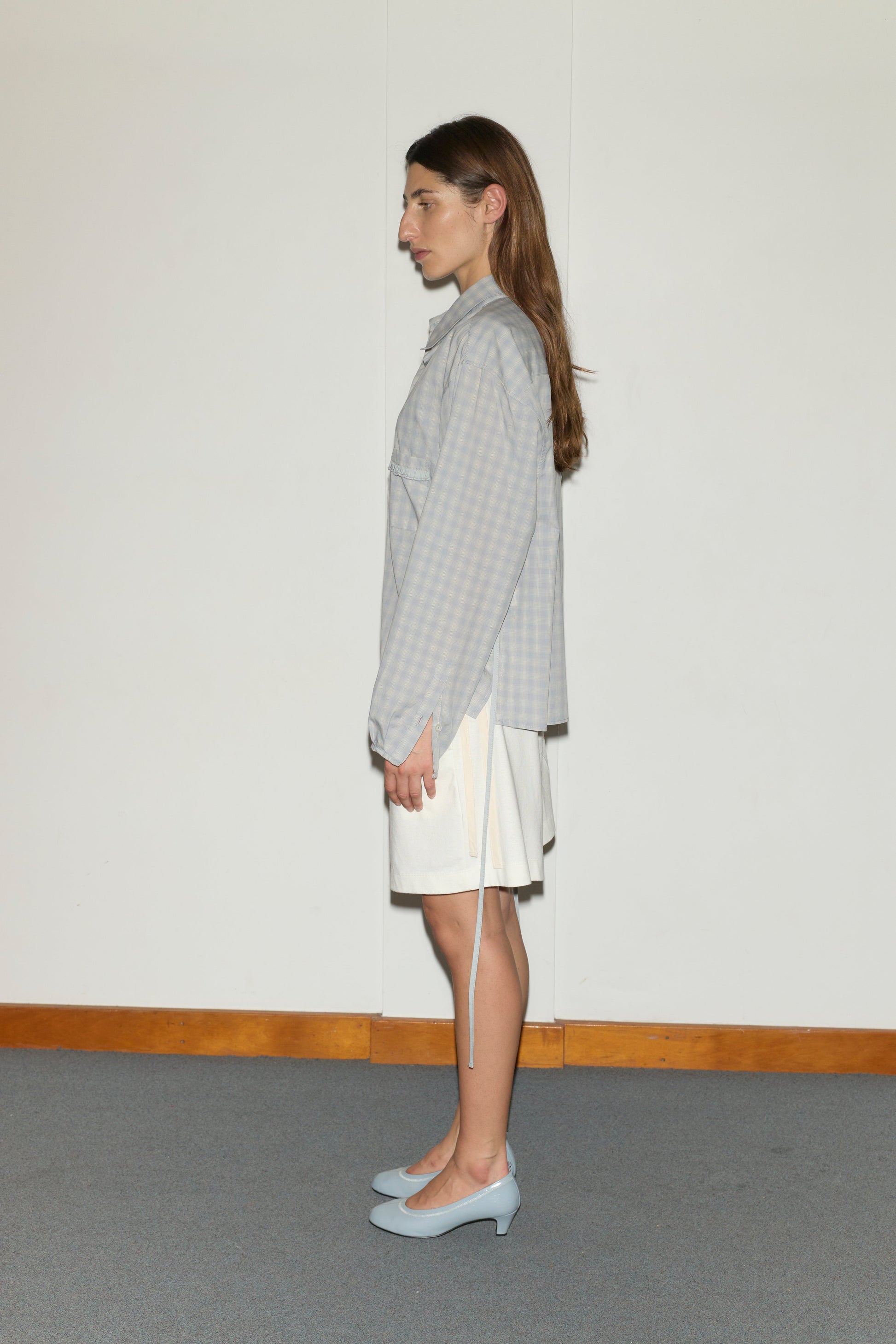 A person with long brown hair stands in profile against a white wall, wearing the Relax Short - Off White set with a white skirt and light blue heels on a gray carpeted floor with wood trim.