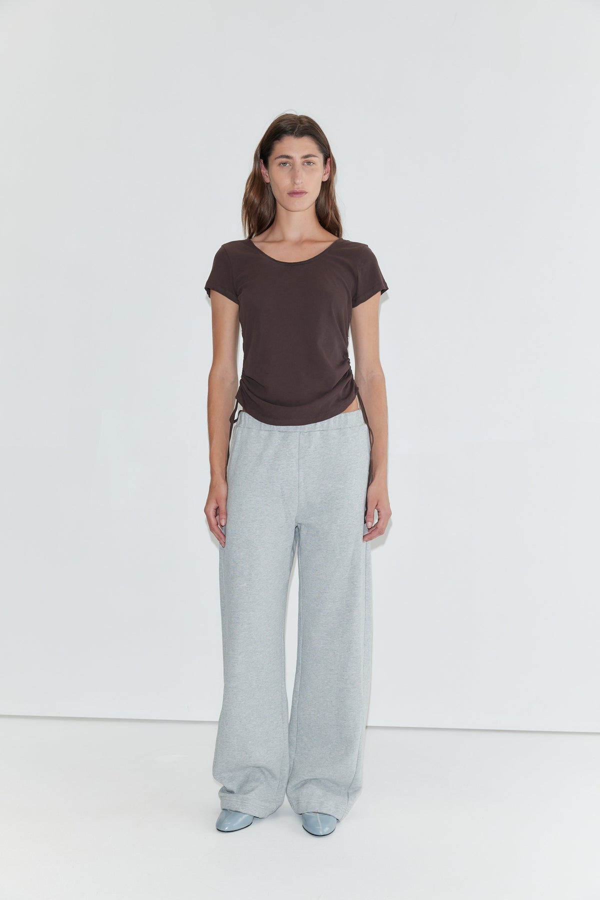 A woman stands against a plain white background, wearing the Draw Side Tee in brown—a relaxed cotton jersey short-sleeve top. She pairs it with loose light gray sweatpants and light blue shoes. Her straight brown hair frames her neutral expression.