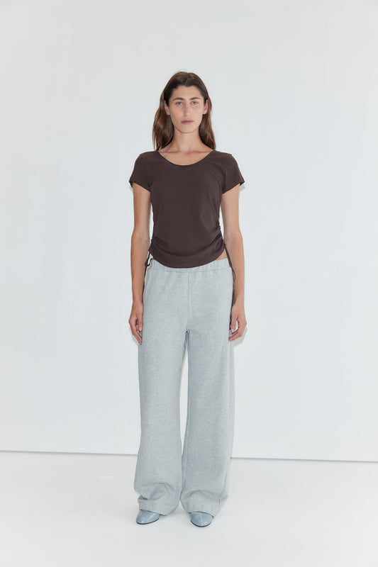 A woman stands against a plain white background, wearing the Draw Side Tee in brown—a relaxed cotton jersey short-sleeve top. She pairs it with loose light gray sweatpants and light blue shoes. Her straight brown hair frames her neutral expression.
