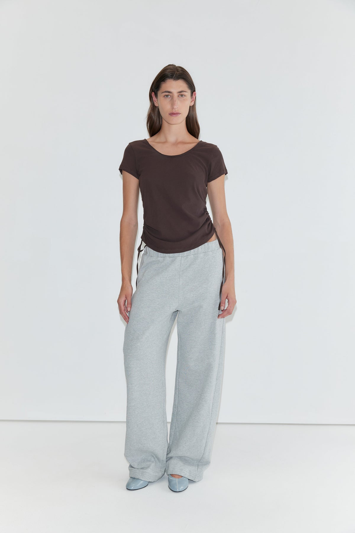 A person stands against a plain white background wearing a brown short-sleeved top, the Side Pleat Pant in Grey Marle—evoking Deiji Studios’ effortless style—and light blue shoes.