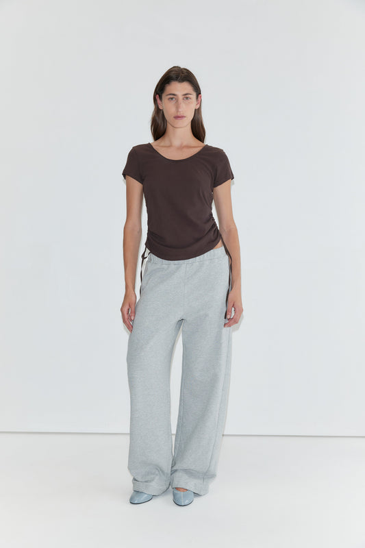 A person stands against a plain white background wearing a brown short-sleeved top, the Side Pleat Pant in Grey Marle—evoking Deiji Studios’ effortless style—and light blue shoes.