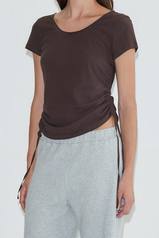 A person wearing the Draw Side Tee - Brown, a short-sleeve ruched cotton jersey top with side ties, paired with light gray sweatpants and standing against a plain white background.