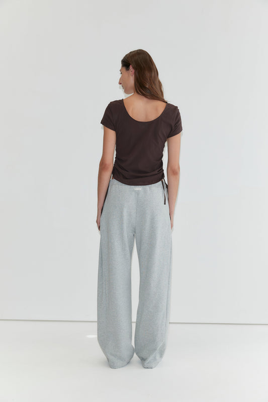 A person with long brown hair stands facing away, wearing the Draw Side Tee - Brown crafted from cotton jersey and loose light gray sweatpants, against a plain white background—a relaxed look reminiscent of deiji studios.