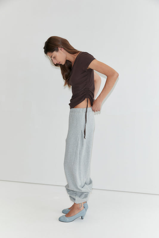 A woman in the Draw Side Tee - Brown by Deiji Studios and loose gray sweatpants, rolled up at the ankles, stands against a white background, looking down as she adjusts her cotton jersey shirt. She wears light blue high-heeled shoes.