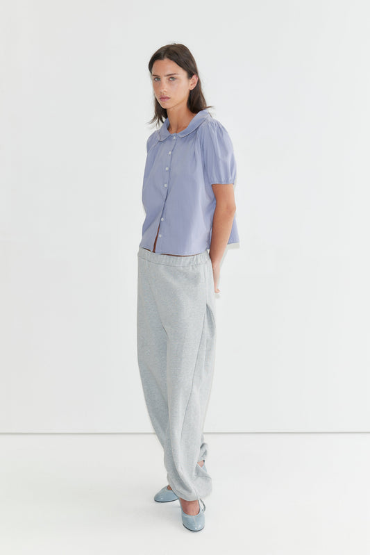 A woman with long dark hair stands against a plain white background wearing the Petal Top - Thistle by Deiji Studios, a pale periwinkle petal-collar blouse, with grey marle trousers and pale blue heels.