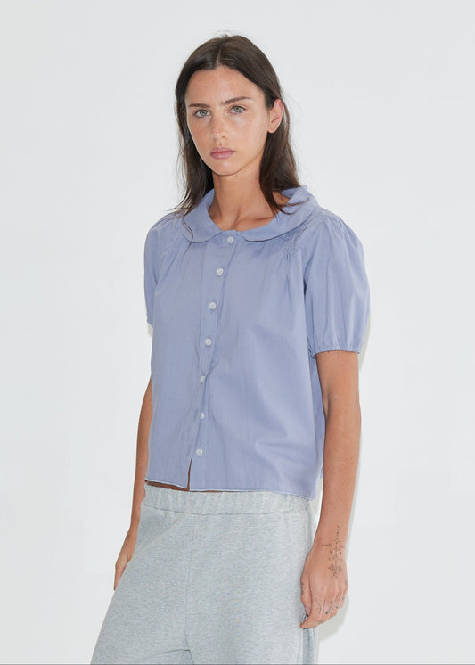 A cropped front view of the Petal Top - Thistle by Deiji Studios, a pale periwinkle short-sleeve blouse with a rounded petal collar, front buttons and puff sleeves, with grey marle trousers against a plain white background.