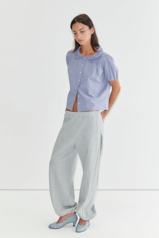 A woman with long dark hair stands against a plain white background looking down, wearing the Petal Top - Thistle by Deiji Studios, a pale periwinkle petal-collar blouse with puff sleeves, and grey marle trousers with pale blue heels.