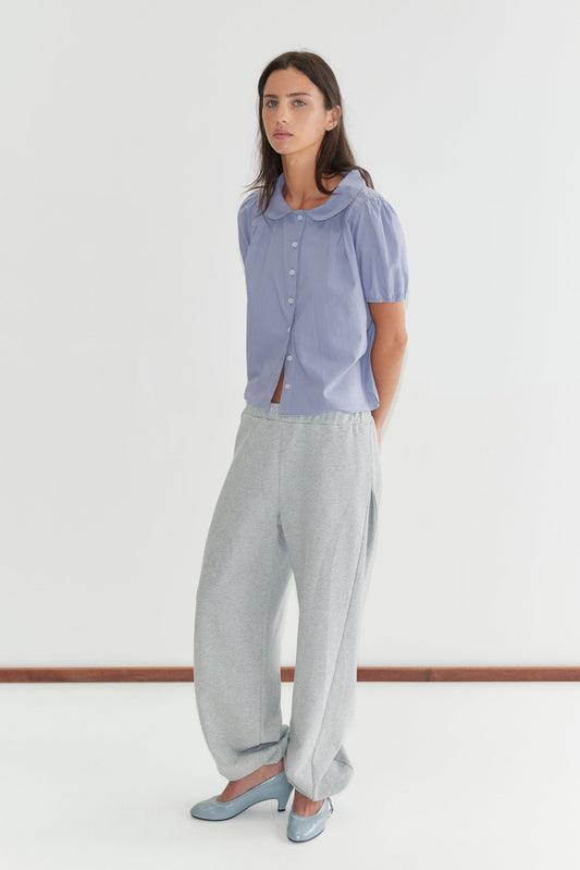 A woman with long dark hair stands against a plain white background wearing the Petal Top - Thistle by Deiji Studios, a pale periwinkle petal-collar blouse with front buttons and puff sleeves, with grey marle trousers and pale blue heels.
