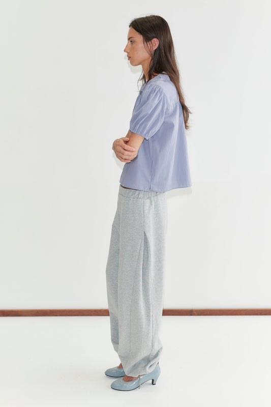 A woman with long dark hair stands in profile against a plain white background wearing the Petal Top - Thistle by Deiji Studios, a pale periwinkle petal-collar blouse, with grey marle trousers and pale blue heels.