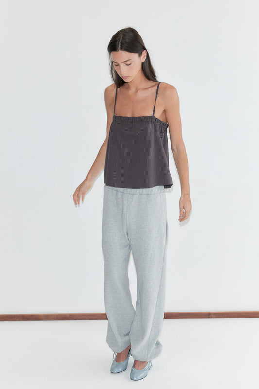 A woman with long dark hair stands against a plain white background looking down, wearing the Frill Placket Top - Brown Blue Stripe by Deiji Studios, a dark brown fine-striped cami top with a frill placket, with grey marle trousers and pale blue heels.